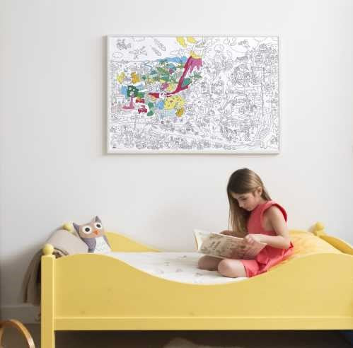 Criança sentada numa cama amarela a ler, enquanto um poster XL OMY "Dinossauros" parcialmente colorido está pendurado na parede branca.