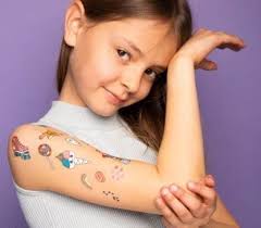 Criança a exibir várias tatuagens temporárias coloridas de unicórnios e doces no braço, com um fundo lilás.