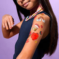 Criança com cabelo comprido a exibir tatuagens temporárias OMY Kawai aplicadas no braço, incluindo um gato vermelho, arco-íris e cogumelo.