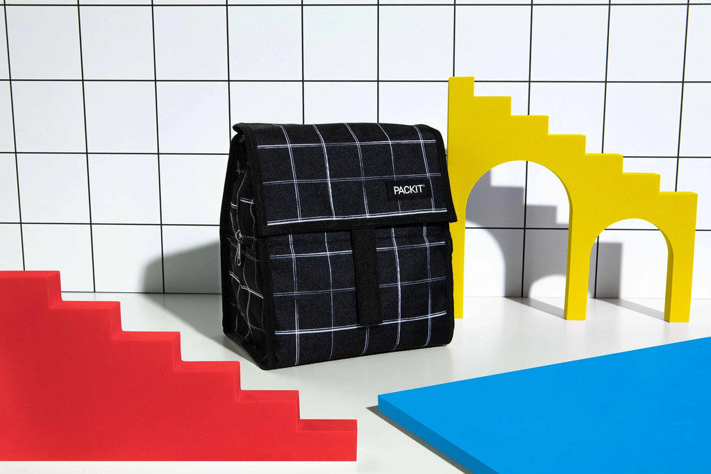 Bolsa lancheira Packit preta com grelha branca em cenário pop art com blocos de escadas amarelos, vermelhos e um plano azul.