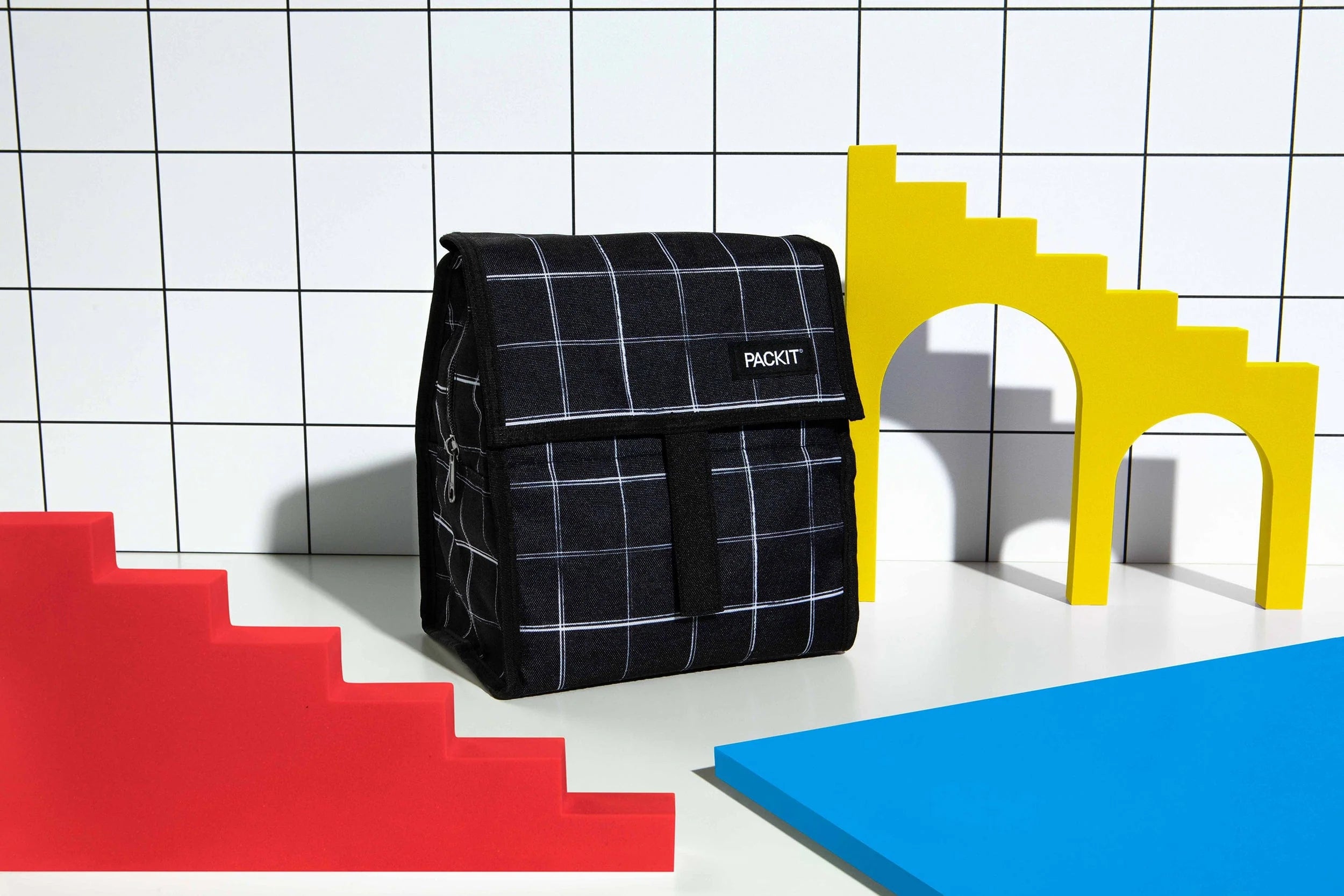 Bolsa lancheira Packit preta com grelha branca em cenário pop art com blocos de escadas amarelos, vermelhos e um plano azul.