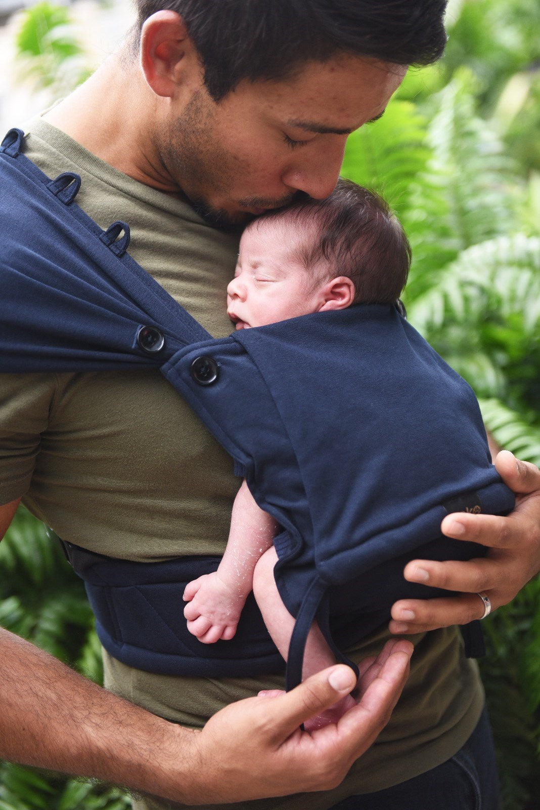 Pai a beijar a testa do bebé recém-nascido que dorme na mochila de babywearing Neko Tiny cor azul marinho, em ambiente exterior.