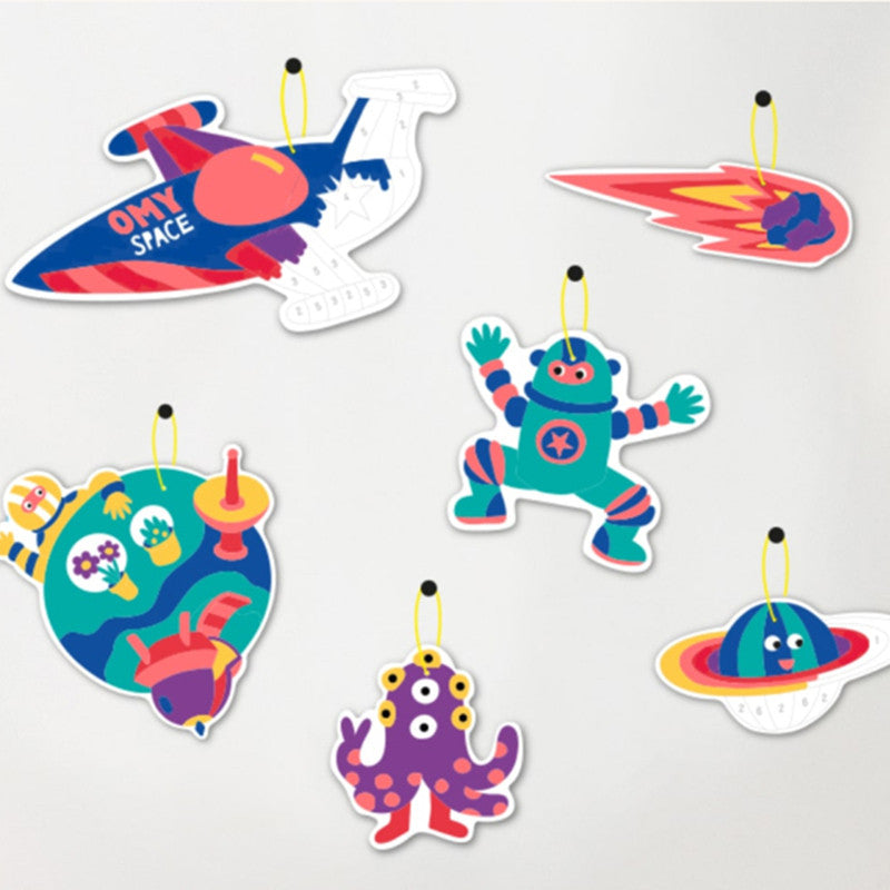 Decorações cósmicas da OMY para colorir, incluindo foguetão, planeta, robô e monstro, prontas para pendurar.