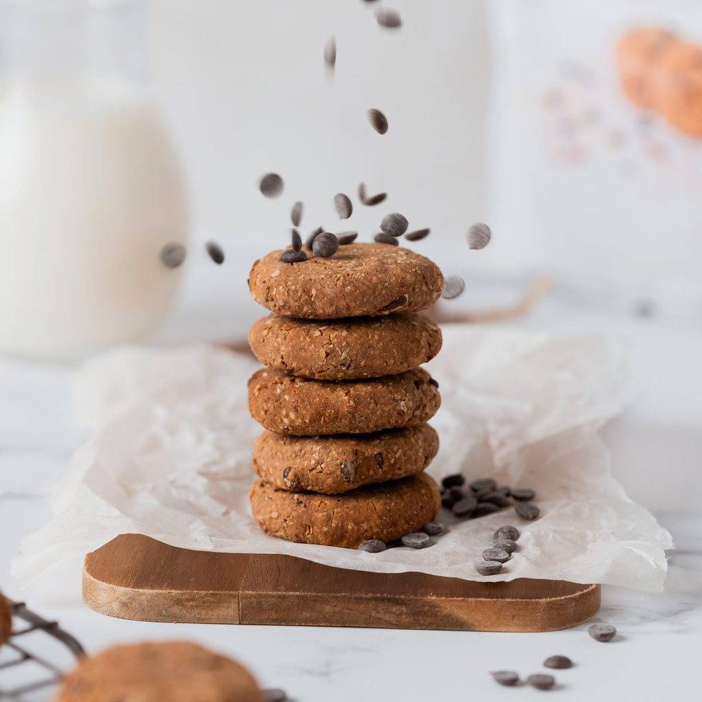 Pilha de cinco bolachas caseiras com pepitas de chocolate, com mais pepitas a cair por cima, sobre uma tábua de madeira e papel vegetal.