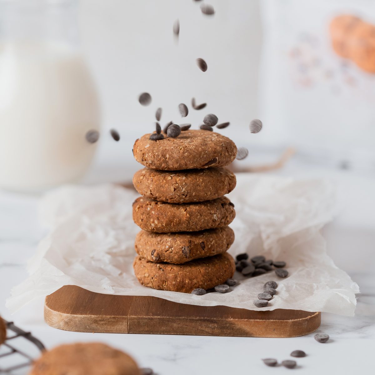 Pilha de cinco bolachas caseiras com pepitas de chocolate, com mais pepitas a cair por cima, sobre uma tábua de madeira e papel vegetal.