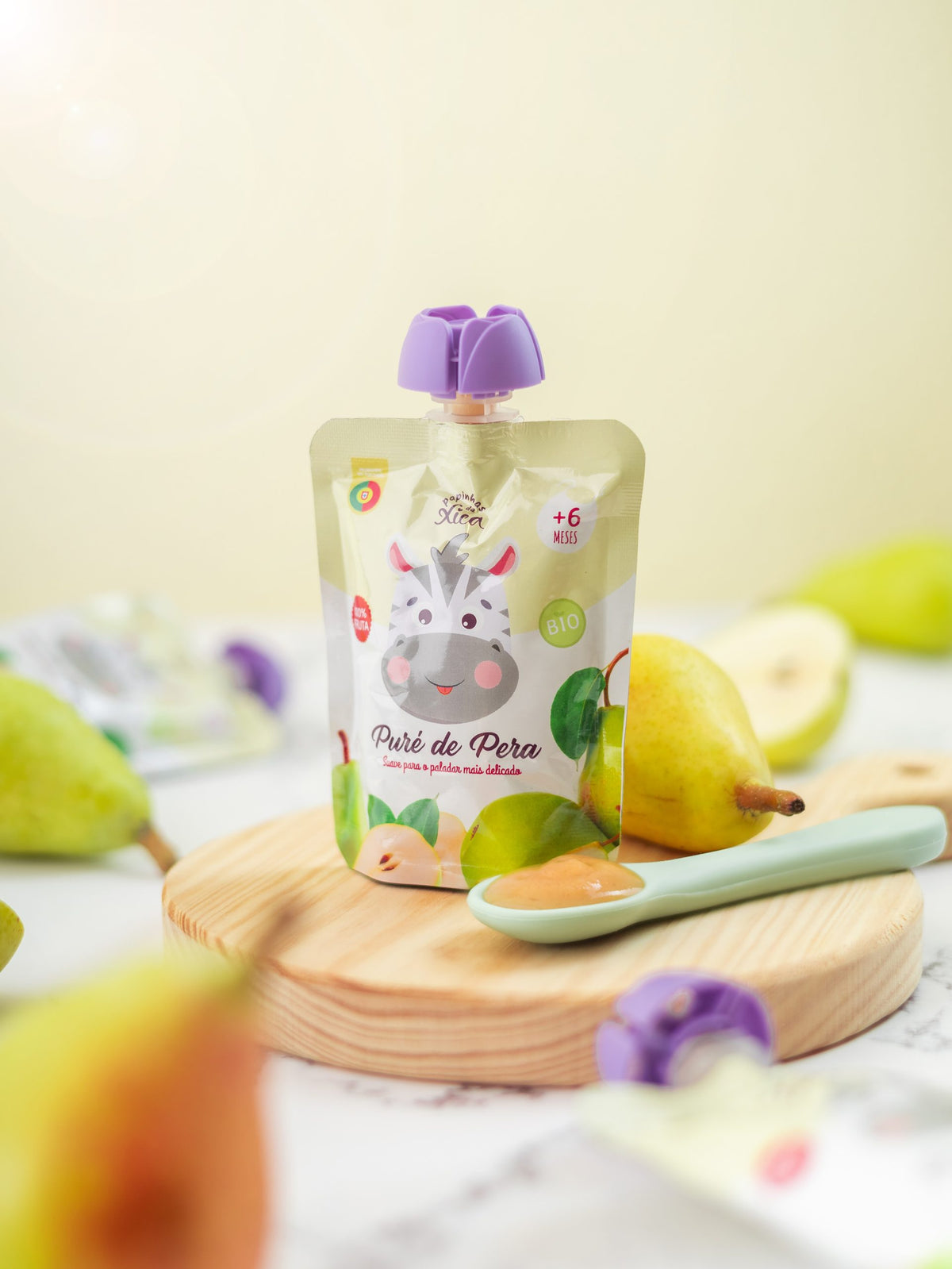 Saqueta de Papinhas da Xica Puré de Pera biológico para bebés, com colher e peras frescas, sobre uma tábua de madeira.
