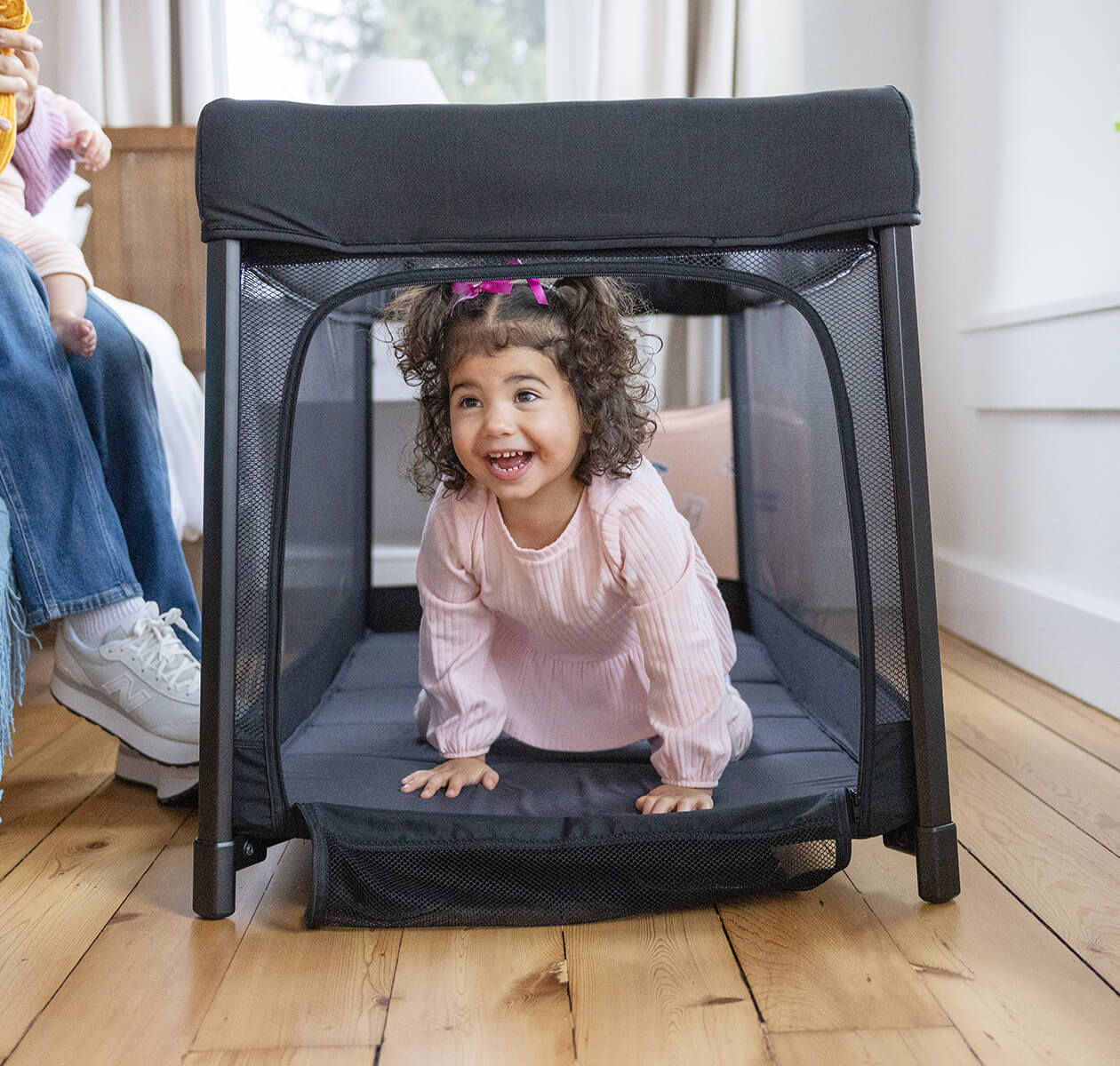 Criança pequena sorridente a gatinhar para fora da entrada lateral com zíper do parque de viagem Joie amigo™ 120.