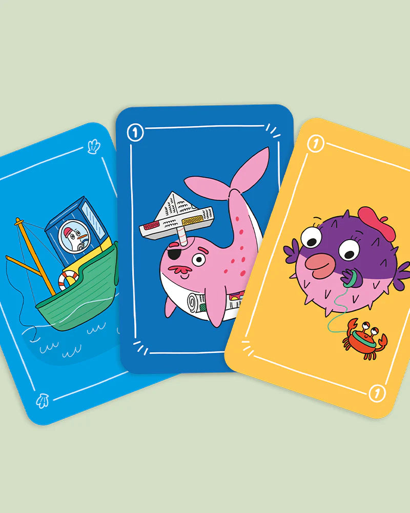 Três cartas do jogo "Vamos Jogar ao Peixinho" com ilustrações cómicas: uma traineira, uma baleia rosa e um peixe-balão roxo com um caranguejo.