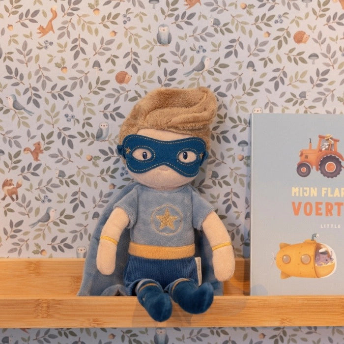 Peluche Super-herói Jim da Little Dutch sentado numa prateleira de madeira, com máscara azul e capa, ao lado de um livro infantil.