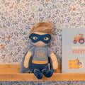 Peluche Super-herói Jim da Little Dutch sentado numa prateleira de madeira, com máscara azul e capa, ao lado de um livro infantil.