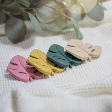 Quatro mordedores de silicone em forma de folha (Monstera) em cores diferentes (rosa, amarelo, verde escuro, bege) dispostos sobre um tecido branco texturado com folhagem desfocada ao fundo.