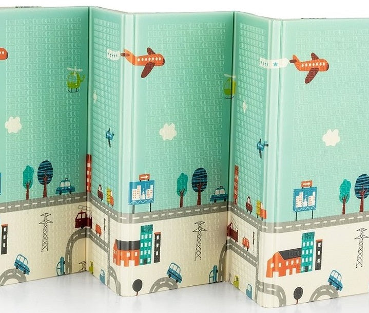 Detalhe do tapete dobrável bbes com o lado "City" aberto, mostrando padrão de cidade, árvores e aviões num fundo azul-esverdeado.