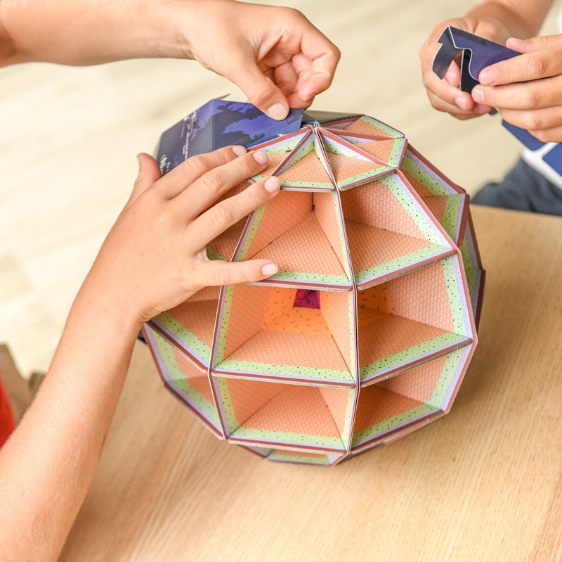 Detalhe da montagem interna do globo 3D Poppik, mostrando as camadas de papelão e as mãos de crianças a encaixar as secções.