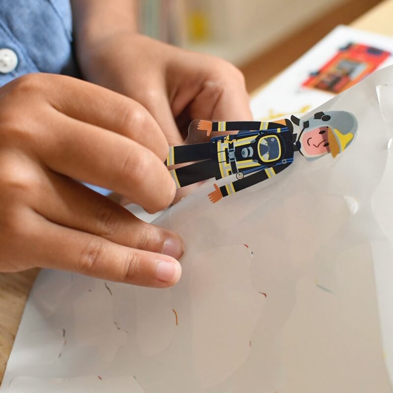 Mãos de criança a retirar um autocolante de bombeiro com equipamento completo, pronto a ser colocado no poster educativo Poppik.