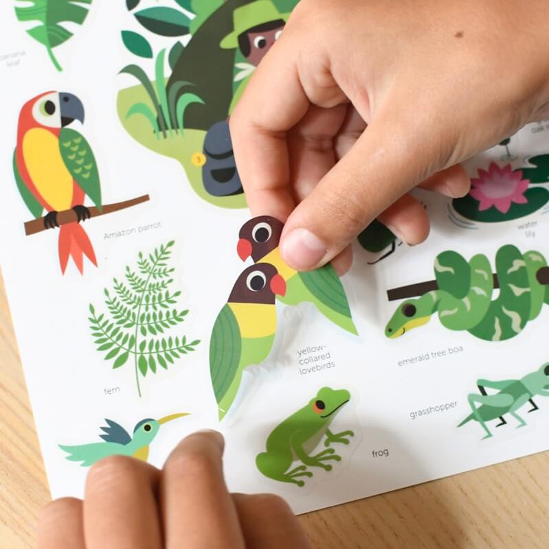 Mãos a aplicar autocolantes de animais da selva, como pássaros e insetos, no poster Poppik Mini DISCOVERY, destacando a atividade de colagem.