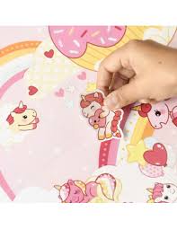 Close-up de uma mão a colar um autocolante de unicórnio fofo num poster de fundo rosa com arco-íris e doces.