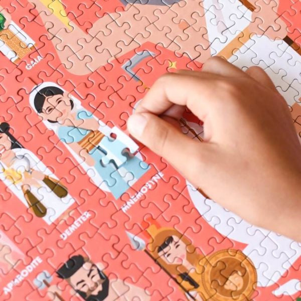 Mão a colocar uma peça do puzzle Poppik Mythology, revelando as figuras de Demeter e Mnemosyne, com cores vibrantes.