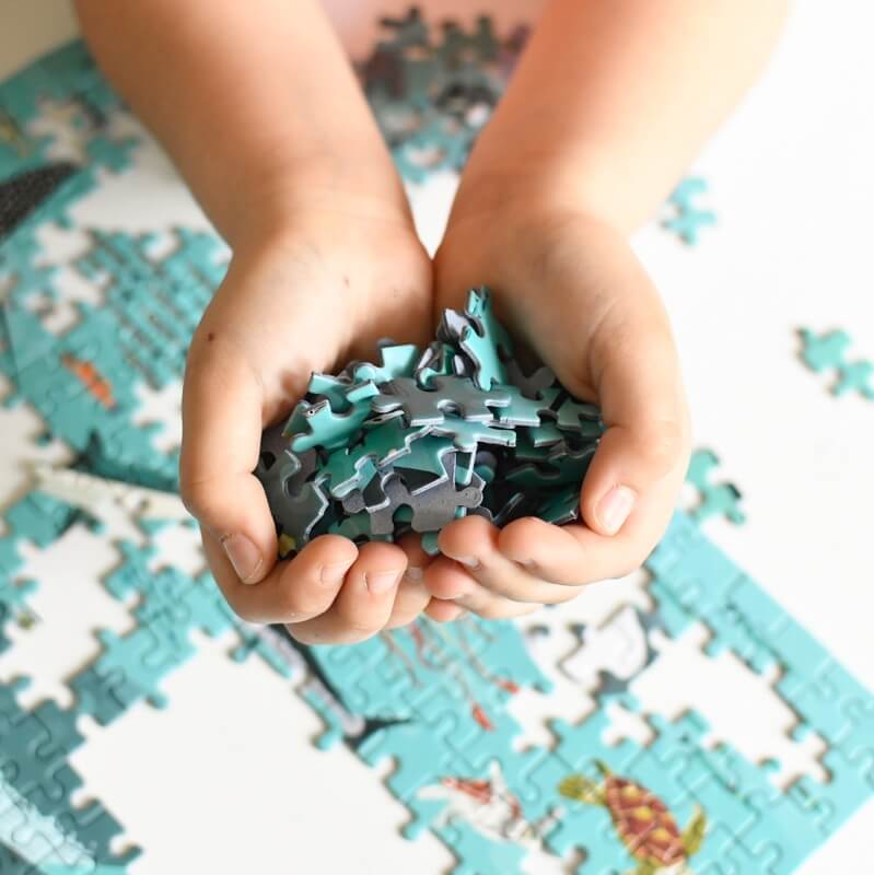 Duas mãos pequenas seguram um punhado de peças do puzzle do oceano Poppik, com o puzzle parcialmente montado visível em primeiro plano.