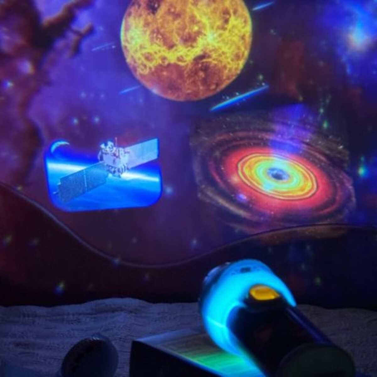 Projeção de espaço com um planeta, galáxia e nave espacial a iluminar um quarto escuro, com a ponta da lanterna azul visível em primeiro plano.