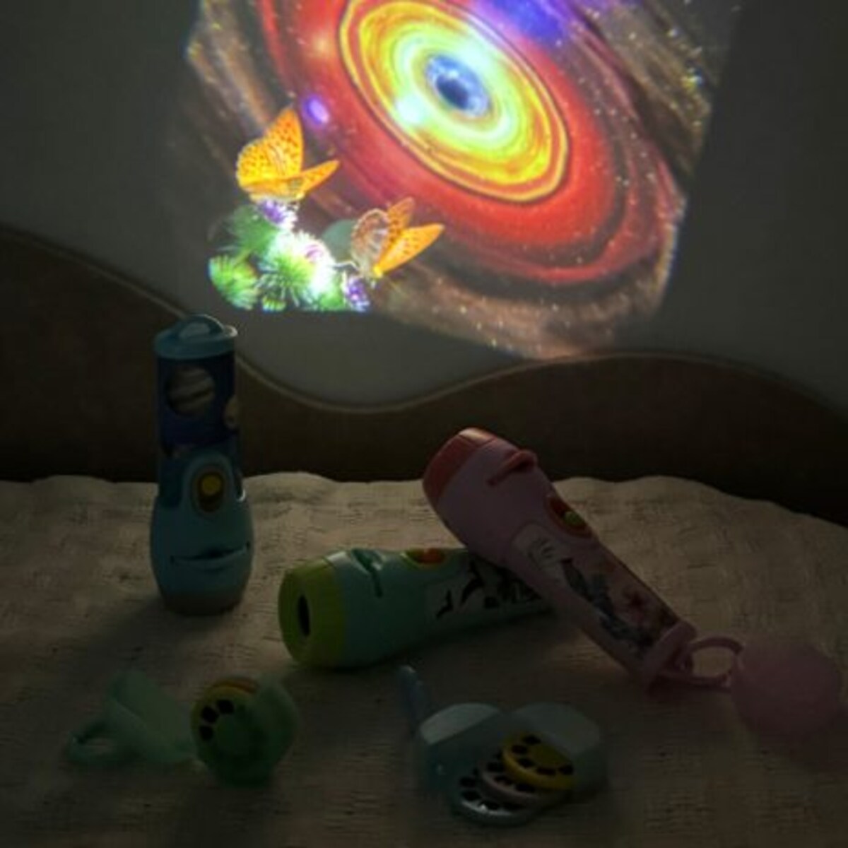Vários projetores de luz (azul, verde, rosa) e discos de projeção espalhados numa cama, com projeções de borboletas e espaço visíveis no fundo.