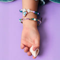 Pulso de uma criança a usar duas pulseiras temáticas de sereia, segurando uma concha natural contra um fundo roxo.