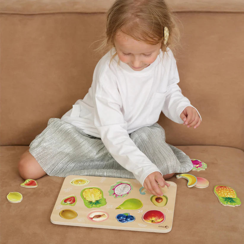 Criança sentada no sofá, a brincar e a encaixar as peças do Classic World Puzzle de encaixe Frutas em madeira, demonstrando a interação com o brinquedo educativo.