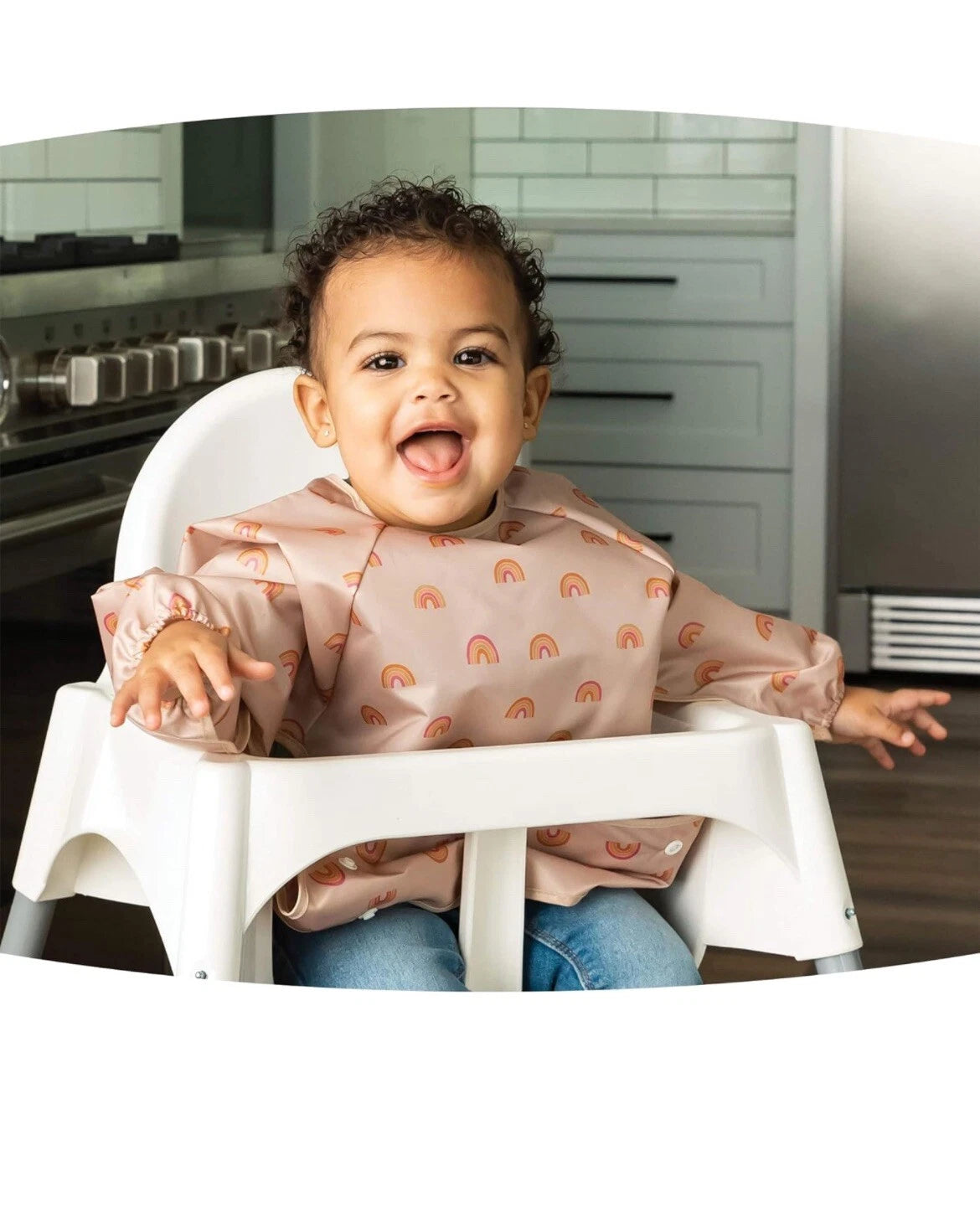 Bebé sorridente a usar o babete impermeável Tiny Twinkle Rainbow com mangas, sentado numa cadeira de refeição na cozinha, pronto para comer.