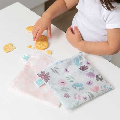 Detalhe dos dois sacos reutilizáveis Bumkins abertos numa mesa branca, enquanto mãos de criança pegam em bolachas, ilustrando o uso para snacks.