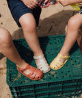 Crianças sentadas numa caixa de plástico verde, usando sandálias Igor Barefoot em várias cores (bege, vermelho e amarelo). Cenário de verão, areia e brincadeiras ao ar livre.