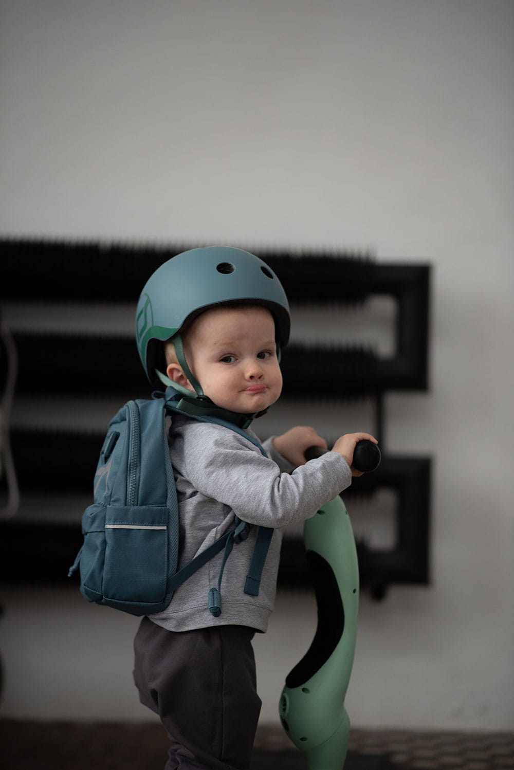 Criança com capacete Scoot & Ride azul 45-51cm (2-5 anos) e mochila, segura trotinete.