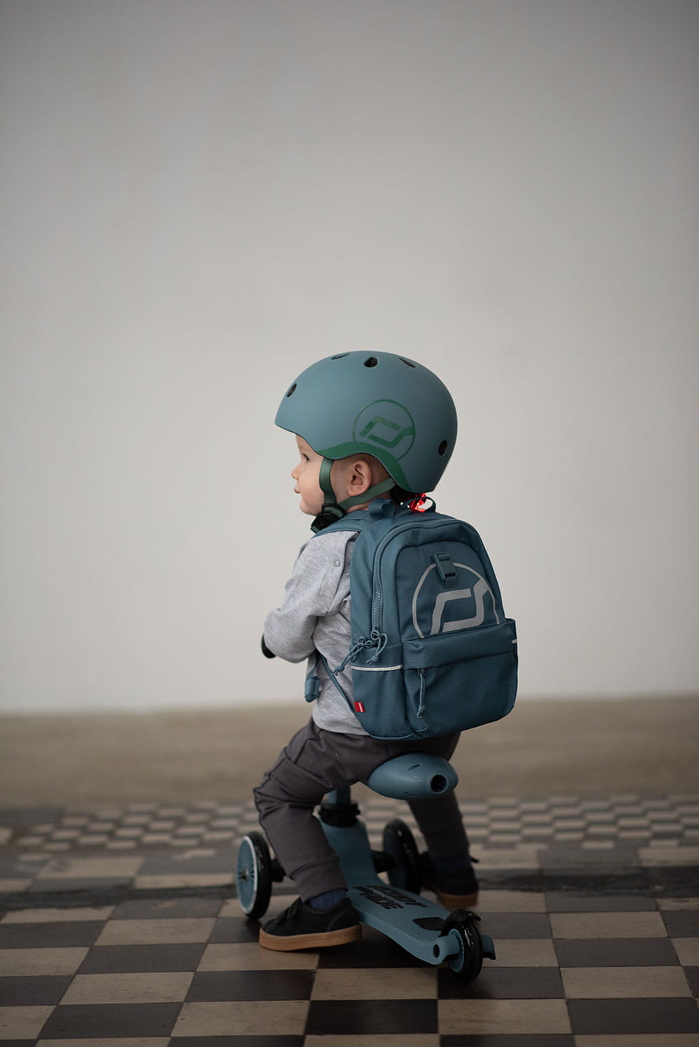 Criança com capacete Scoot & Ride azul 45-51cm (2-5 anos) e mochila, sentada em trotinete.