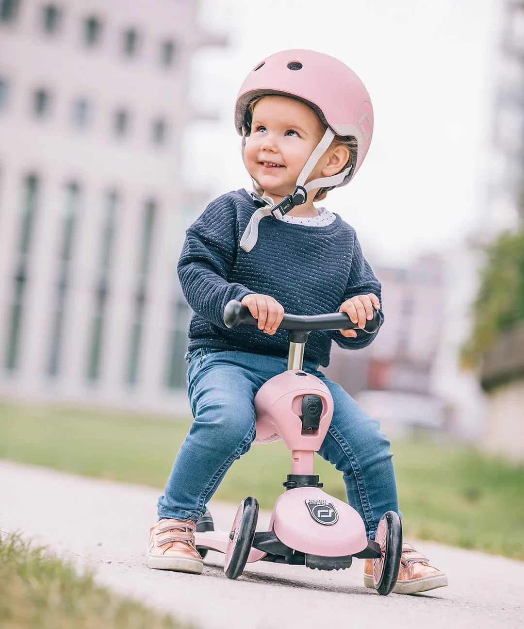 Criança pequena com capacete Scoot & Ride rosa 45-51cm (2-5 anos) sentada em trotinete.