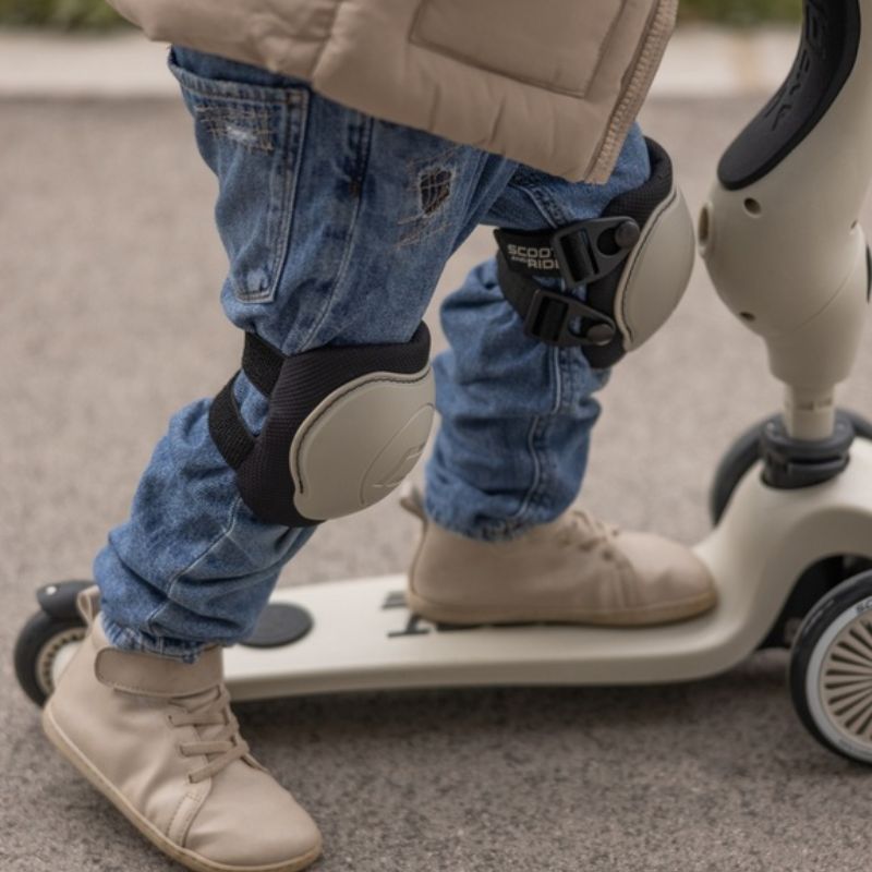 Criança a usar proteções Scoot & Ride bege nos joelhos enquanto anda de trotinete, focando o ajuste seguro.