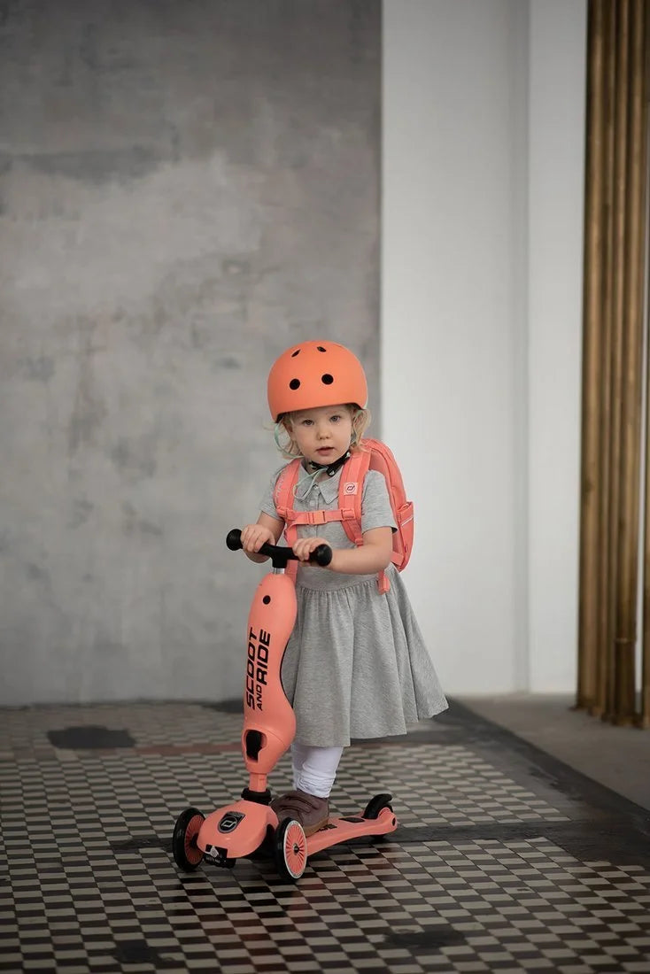 Criança pequena com capacete cor de laranja a usar a Trotinete Highwaykick 1 no modo trotinete, em interior.