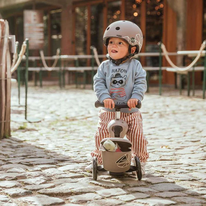 Criança pequena com capacete a usar a Trotinete Highwaykick 1 Lifestyle em modo triciclo, com brinquedo no cesto frontal.