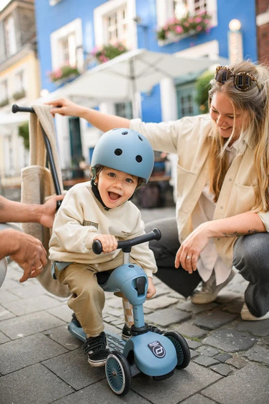 Criança feliz com capacete azul a usar a trotinete Scoot & Ride em modo sentado, com uma mulher a supervisionar em ambiente urbano.