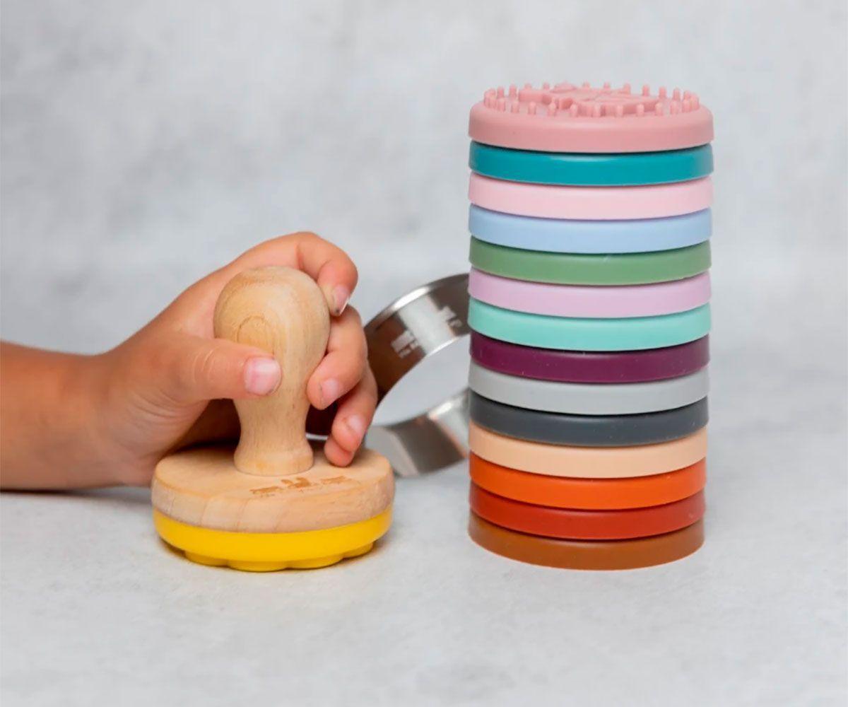 Mão de criança a usar o carimbo de bolachas Monneka com cabo de madeira, com a pilha de carimbos de silicone e o cortador ao fundo.