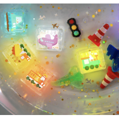 Vista aérea dos 6 Cubos de Luz GloPals AUTO a brilhar intensamente em água, rodeados por confetes e pequenos brinquedos de trânsito.