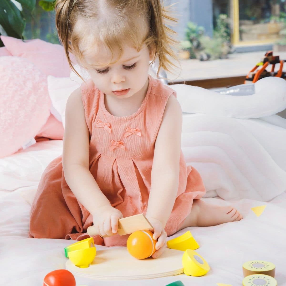 Criança pequena com vestido cor de rosa a brincar com o Classic World Conjunto de frutas para cortar, cortando uma laranja na tábua.