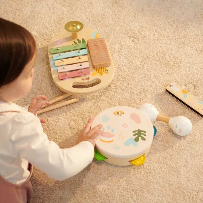 Criança a brincar com o Classic World Set Musical Iris no chão, com a pandeireta nas mãos e o xilofone e maraca por perto.