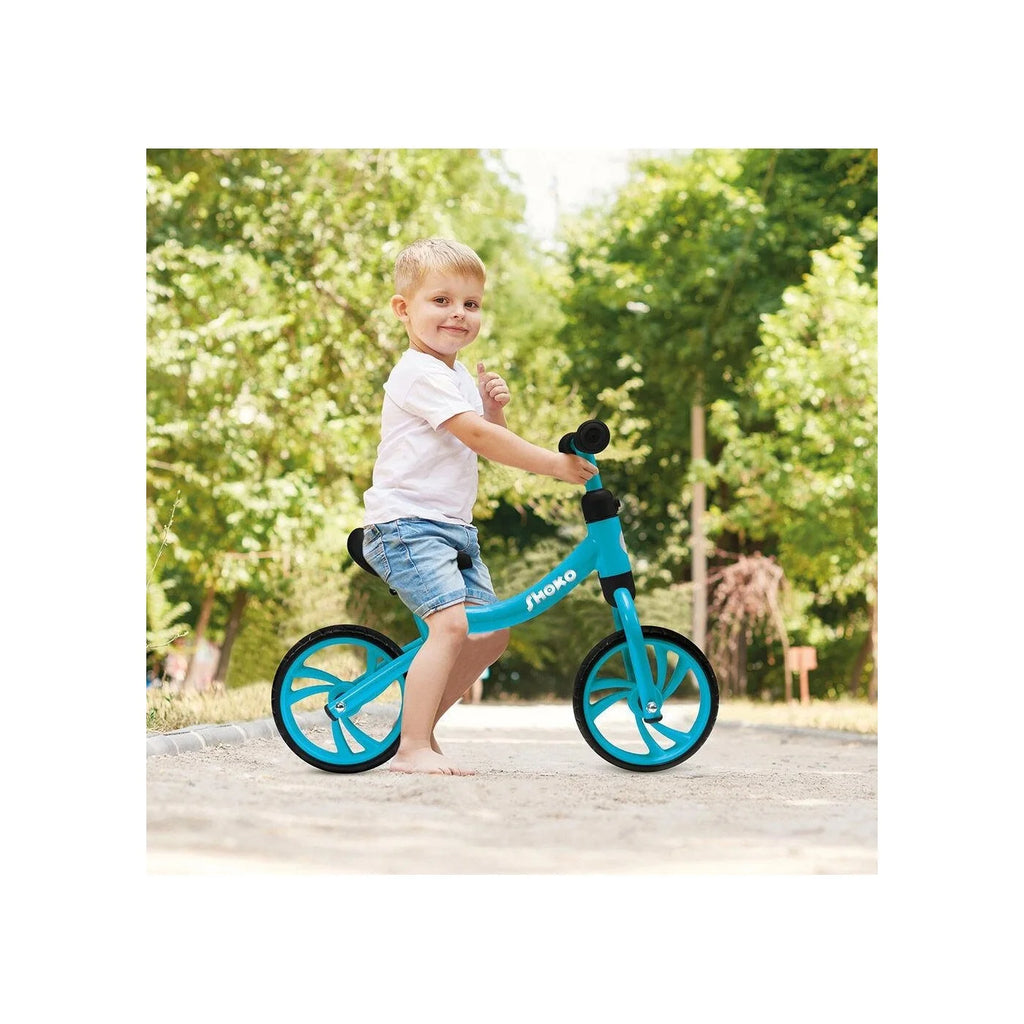 Criança pequena sorridente a andar de bicicleta de equilíbrio Shoko azul num caminho de terra, demonstrando facilidade de uso.