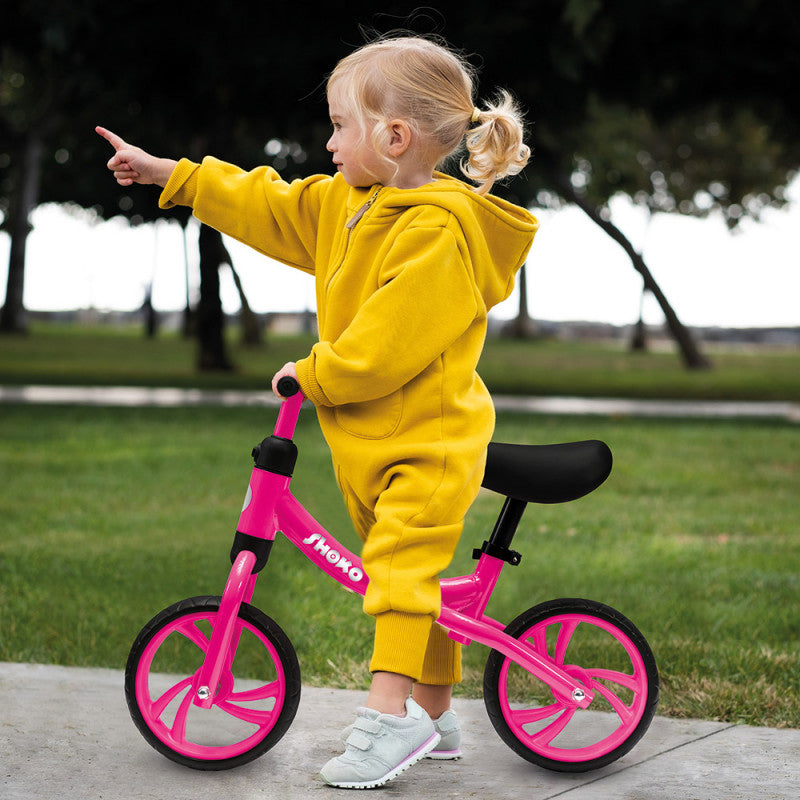 Criança pequena de macacão amarelo a aprender a andar na bicicleta de equilíbrio Shoko rosa num parque, apontando para a frente.