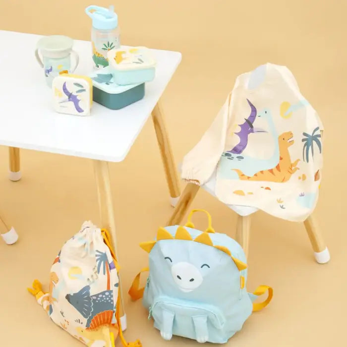 Conjunto de artigos Miniland da coleção Dinossauros, incluindo o babete com mangas, mochila e lancheira, dispostos numa mesa infantil.