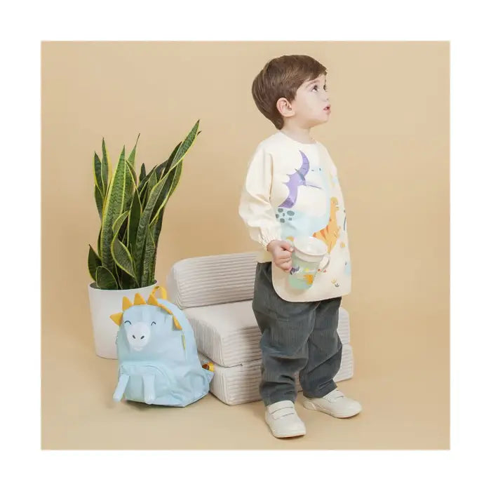 Criança a usar o Miniland babete com mangas Dinossauros, em pé, com mochila a condizer ao lado, num ambiente infantil.