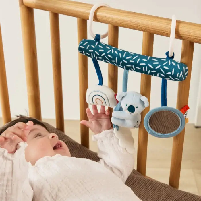 Bebé deitado no berço, a olhar e a interagir com a Miniland Espiral do Coala pendurada, demonstrando a sua utilização.