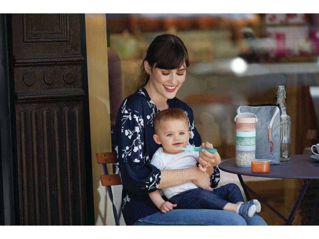Mãe sorridente a alimentar o bebé com uma colher, ao lado da mesa onde está o porta-snacks Skip Hop empilhado dentro do saco de transporte.