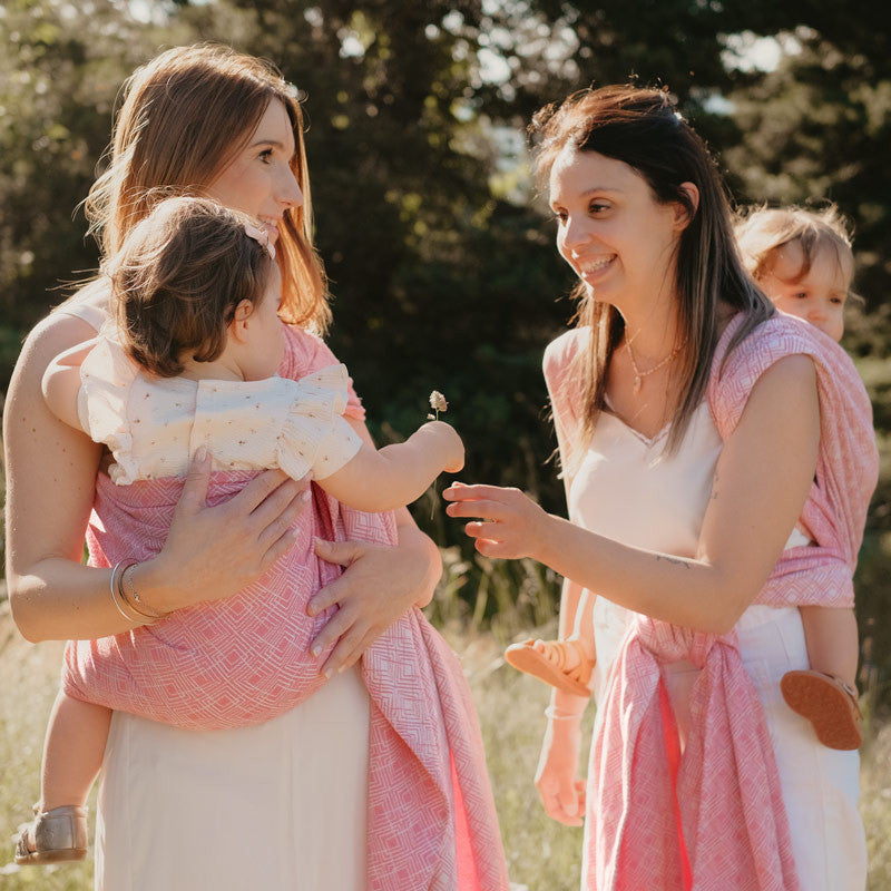 Duas mães ao ar livre, uma a carregar um bebé no sling cor-de-rosa enquanto a outra ajuda a ajustar.