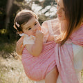 Mãe sorridente a carregar o seu bebé no sling cor-de-rosa, em posição lateral, ao ar livre.