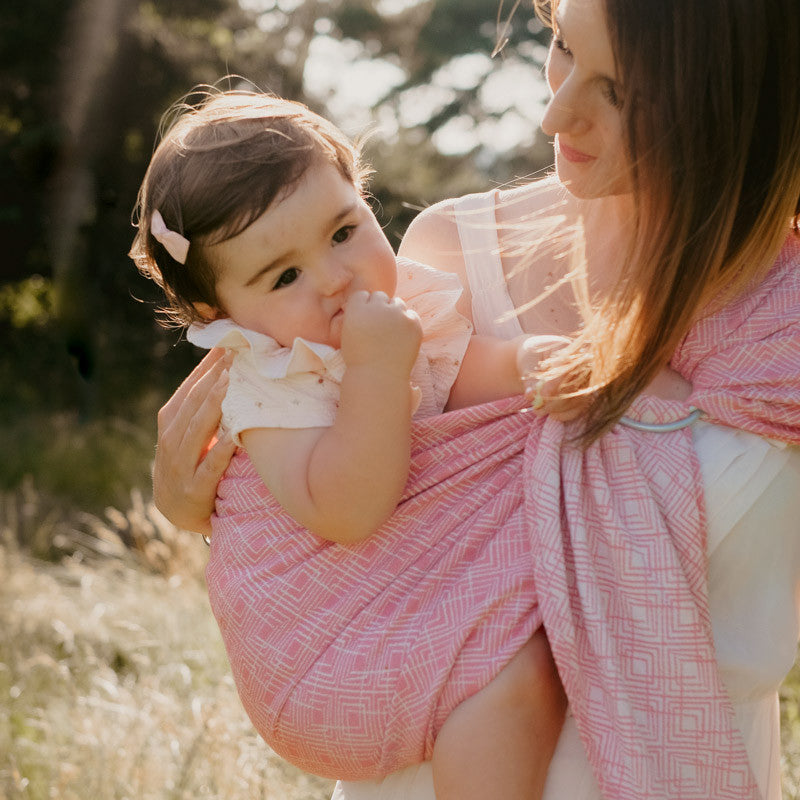 Mãe sorridente a carregar o seu bebé no sling cor-de-rosa, em posição lateral, ao ar livre.