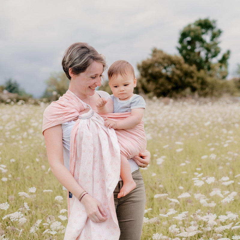 Mãe utilizando o Sling de Argolas Néobulle JACQUARD Ginkgo Rose para carregar o seu bebé num campo florido.